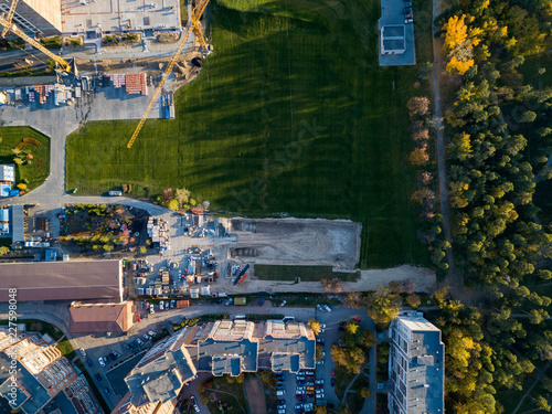 Aerial view of a building construction site at the storage of building materials surrounded by a green lawn with grass at sunset on an autumn sunny day on the border with forest and green trees