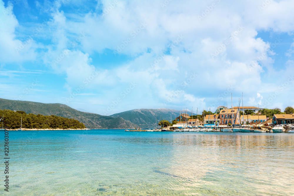 Bright houses in Grecian village Fiscardo, Kefalonia with turquoise sea and cloudy sky above.