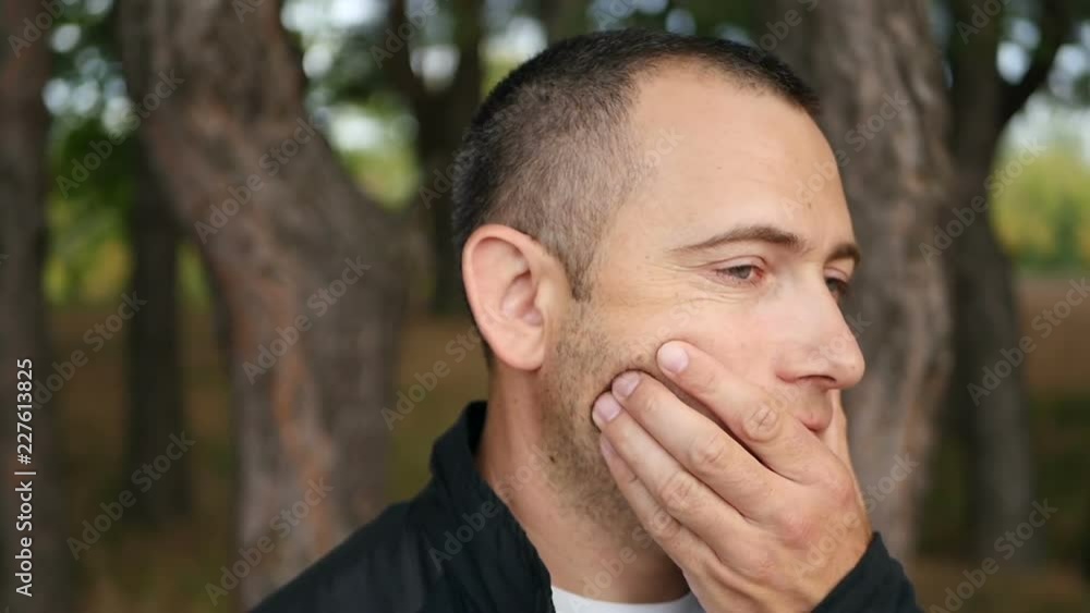 A pensive male tourist is standing outdoors in the forest. Handsome, brutal man touches his face with stubble.