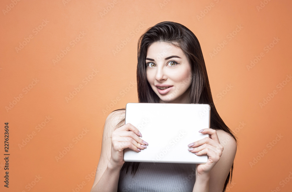 Closeup portrait of a young excited brunette girl with tablet in her hands looking at the camera with open mouth against orange background. Lifestyle, people and technology concept Closeup portrait of a young excited brunette girl with tablet in her hands looking at the camera with open mouth against orange background. Lifestyle, people and technology concept