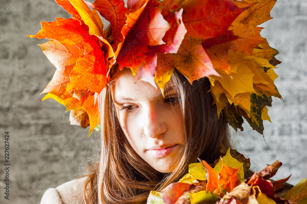 portrait of young teenager brunette girl with long hair with a wreath of autumn leaves on her head on gray wall background