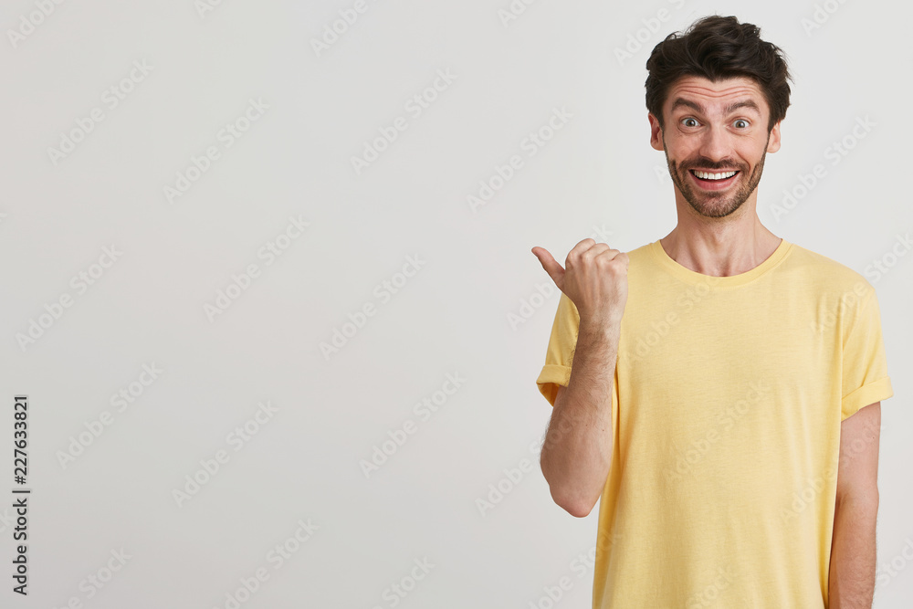 Studio shot of smiling happy attractive young man with dark brown hair and beard wears casual yellow tshirt, points left with one thumb, standing on the right isolated over white background Studio shot of smiling happy attractive young man with dark brown hair and beard wears casual yellow tshirt, points left with one thumb, standing on the right isolated over white background