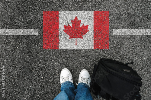 a man with a shoes and backpack is standing on asphalt nex to flag of Canada and border