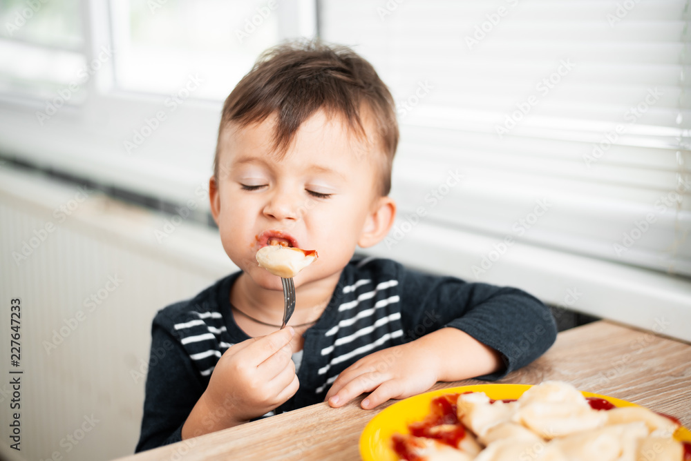 Hungry child eating dumplings in the kitchen, sitting at the table in a gray jacket Hungry child eating dumplings in the kitchen, sitting at the table in a gray jacket