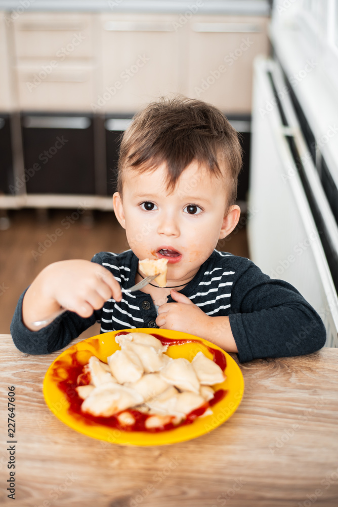 Hungry child eating dumplings in the kitchen, sitting at the table in a gray jacket Hungry child eating dumplings in the kitchen, sitting at the table in a gray jacket
