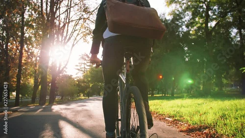 Young businessman with leather bag riding bicycle to city park, slow motion, back view photo