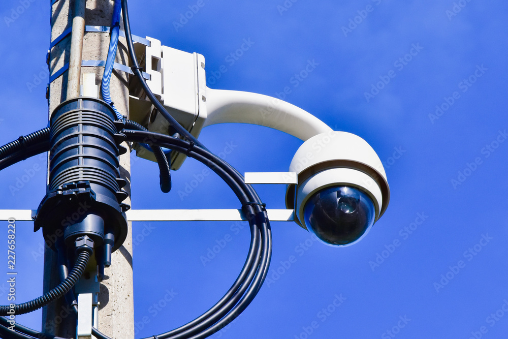 A round-shaped video surveillance camera mounted on a lighting post. Equipment on a blue sky background A round-shaped video surveillance camera mounted on a lighting post. Equipment on a blue sky background
