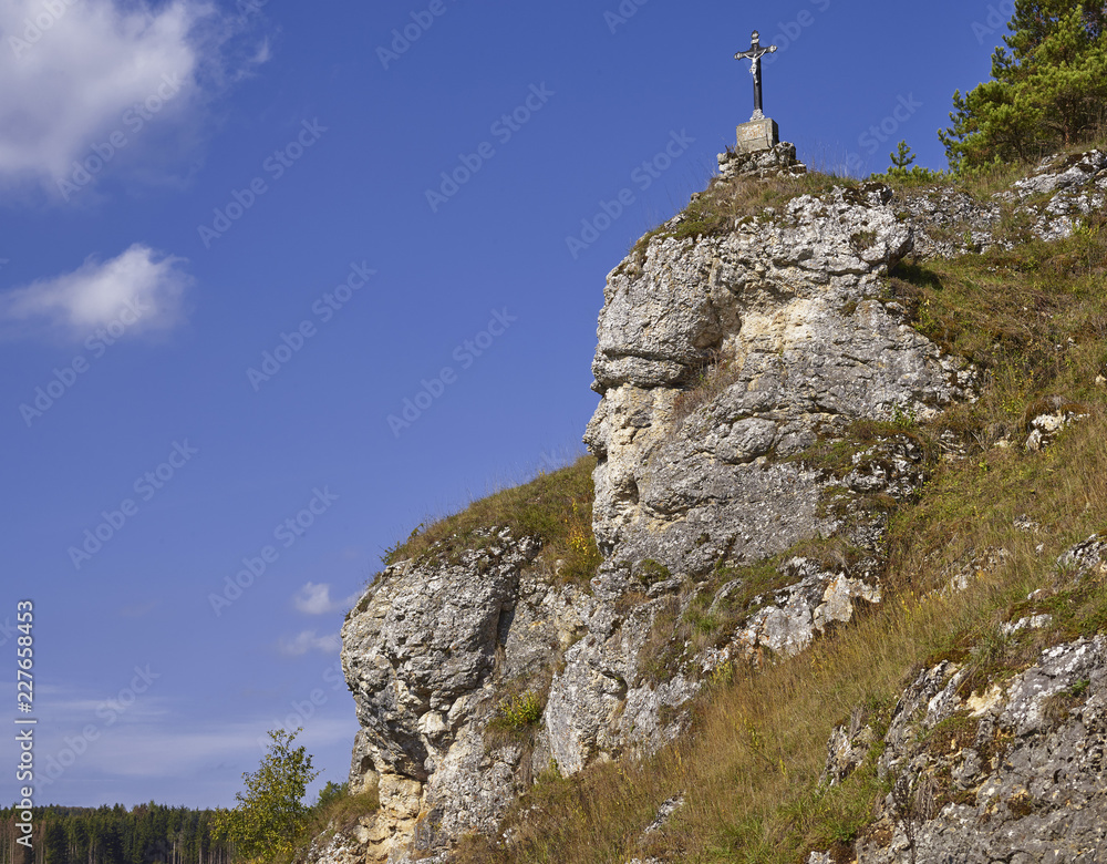 Felswand mit Gipfelkreuz und grüner Bergwiese vor blauem Sommerhimmel