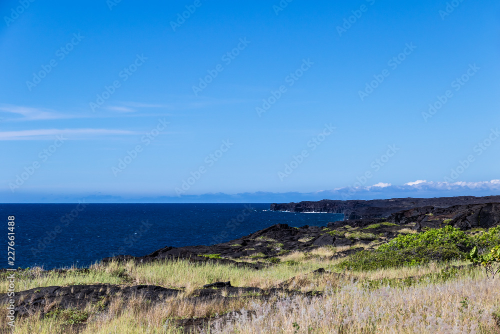 View of rugged coastline on Hawaii's Big Island in Volcano National Park. Black volcanic rocks and vegetation are in the foreground; deep blue Pacific ocean and sky are in the distance. 
