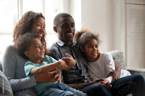 Whole black African funny joyful family sitting together on couch in living room at home. Cheerful married couple little toddler son preschool daughter watching television comedy laughing feels happy
