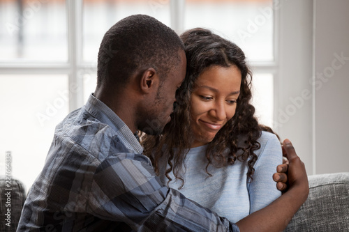 Close up black African married couple in love sitting on couch at home. Loving husband embrace beloved wife after quarrel. Loving boyfriend bored girlfriend. Break up, boredom in relationship concept