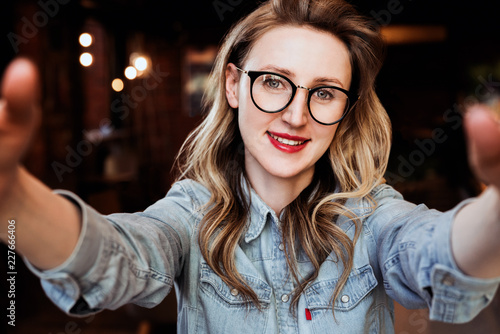 Selfie portrait of young smiling woman sitting in cafe. Hipster girl in trendy glasses takes a selfie in coffee shop.
