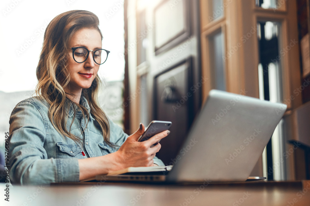 Young businesswoman is sitting in coffee shop at table in front of computer and notebook,using smartphone. Social media. Young businesswoman is sitting in coffee shop at table in front of computer and notebook,using smartphone. Social media.