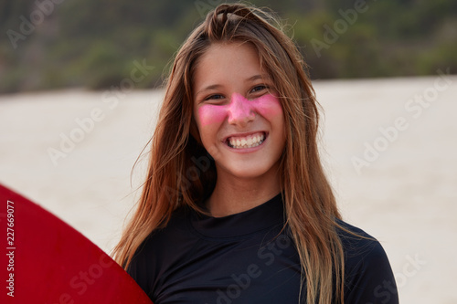 Headshot of cheerful young tourist learn how to surf, poses against sea shore background, has pleased expression, shows white teeth, satisfied with ride on waves. Protection from sun on face photo