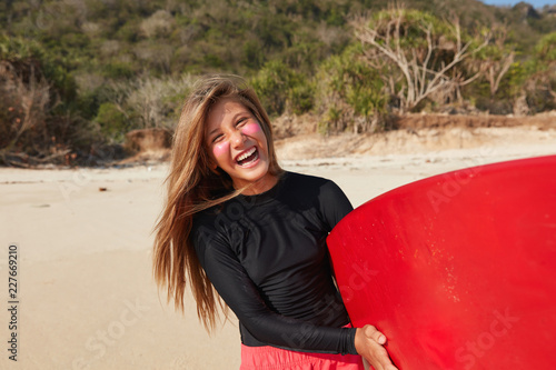 Paddleboarding and wakesurfing concept. Cheerful long haired female fond of surfing, ready to catch ocean and glide across water until wave breaks its energy, poses against sandy beach with board photo