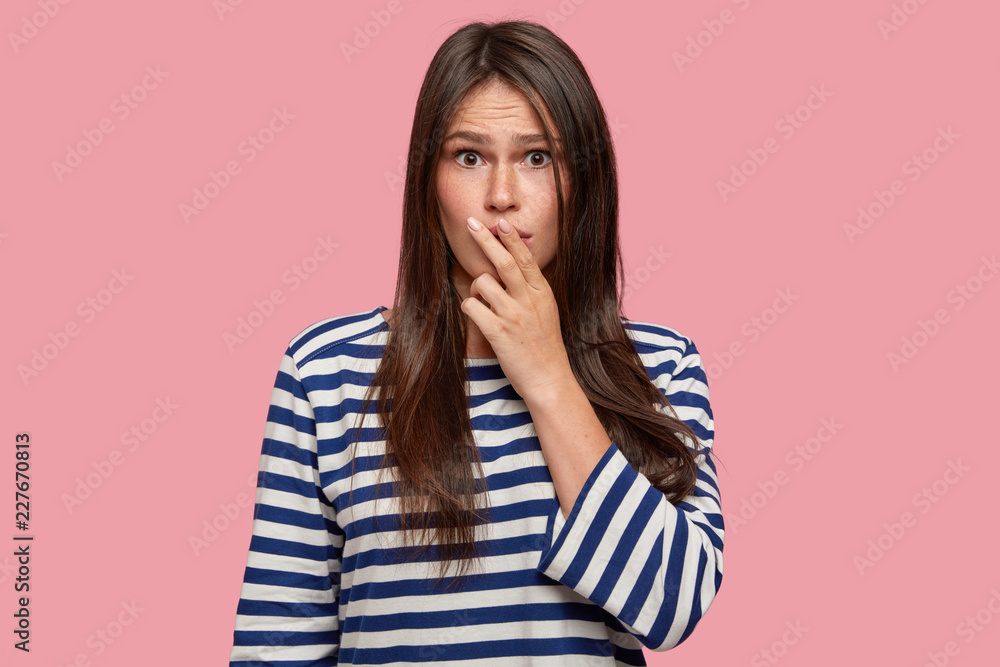 Studio shot of puzzled worried girl keeps both fingers on lips, has frustrared gaze at camera, tries to find solution, being cheated by swindler, models against pink background, dressed casually Studio shot of puzzled worried girl keeps both fingers on lips, has frustrared gaze at camera, tries to find solution, being cheated by swindler, models against pink background, dressed casually