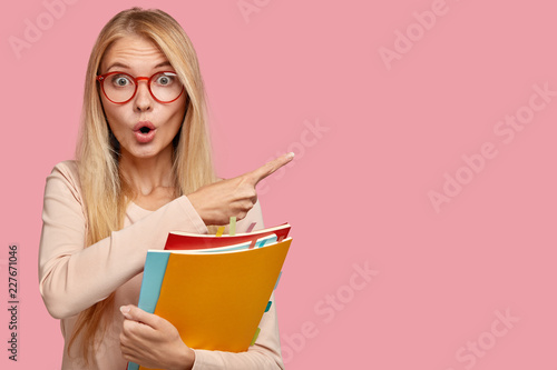Photo of surprised teacher indicates aside with index finger, has light straight hair, keeps mouth opened with amazement, carries textbooks, isolated on pink background. Omg, look at this space photo