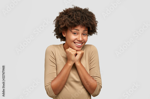 Positive black girlfriend recieves good comments in her blog  keeps both hands under chin  feels pleasure  isolated over white background. African American woman rejoices pleasant moment in life