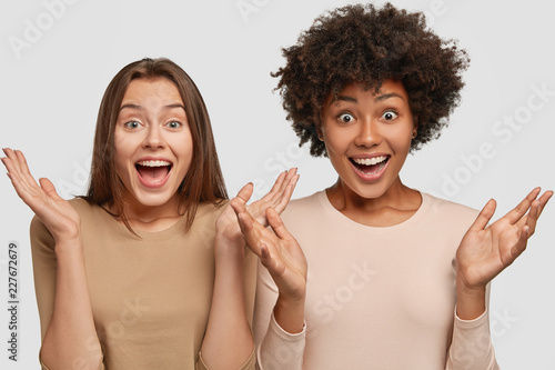 Photo of emotive females gesture with palms, react to awesome news, meet foreign friend, isolated over white background. Amazed dark skinned young woman has Afro haircut spends free time with fellow