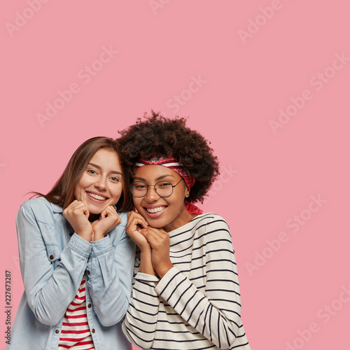 Vertical indoor shot of pleasant looking cheerful mixed race sisters feel support from each other, keep hands together under chin, giggle and stand closely to each other, isolated on pink wall