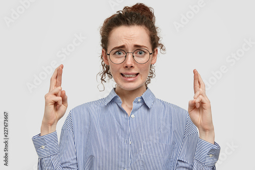 Wallpaper Mural Indoor shot of freckled worried student raise hands with crossed fingers, frowns eyebrows, dressed in fashionable clothes, isolated over white background. Superstitious girl believes in good luck Torontodigital.ca
