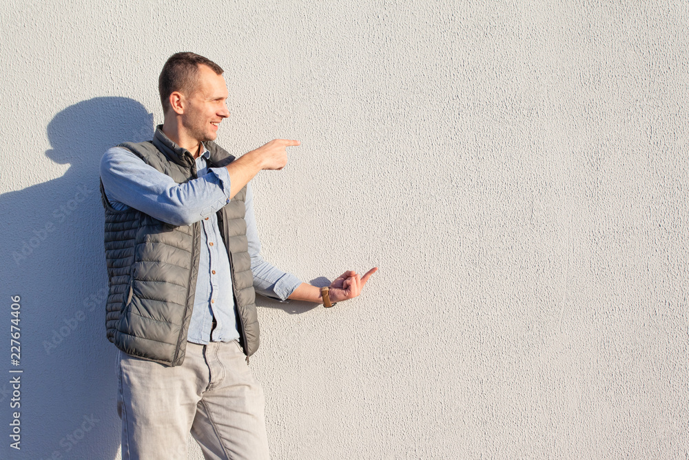 handsome young smiling guy in gray jeans, blue denim shirt and waistcoat stands near the bright wall, copy space