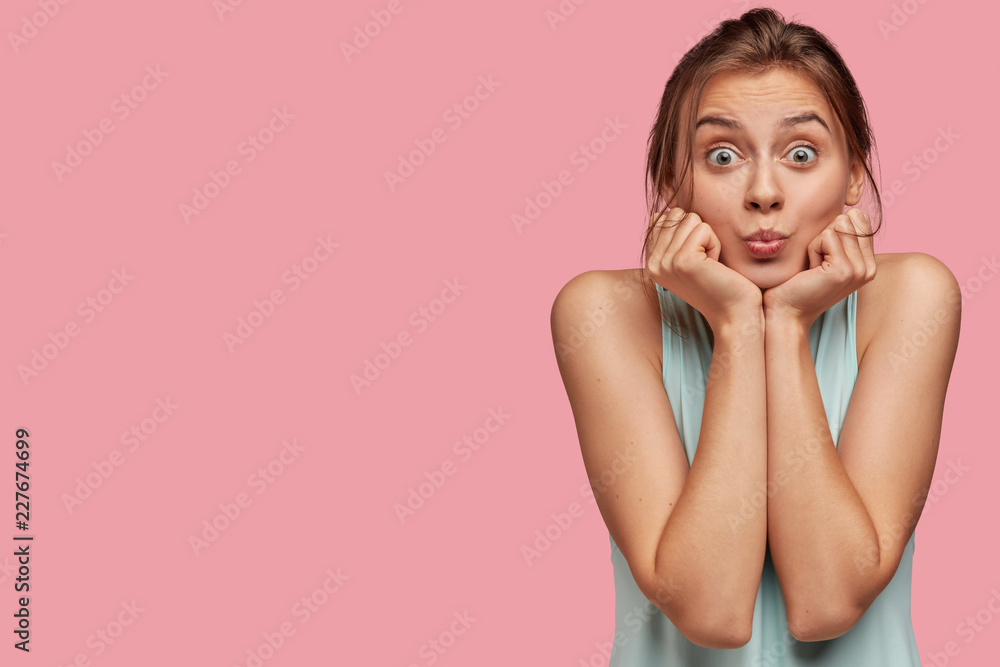 Horizontal shot of beautiful dark haired woman keeps hands under chin and purses lips for making kiss, has stunned expression and bugged eyes, poses over pink background with free space aside on left Horizontal shot of beautiful dark haired woman keeps hands under chin and purses lips for making kiss, has stunned expression and bugged eyes, poses over pink background with free space aside on left