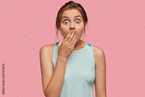 Shocked green eyed beautiful young woman with amazed look, covers mouth with hand, being speechless, dressed in casual clothes, isolated over pink studio background. People and reaction concept.