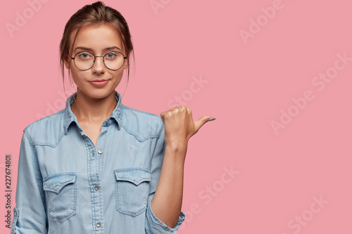 Waist up shot of pleasant looking woman in eyewear, points with thumb at free...