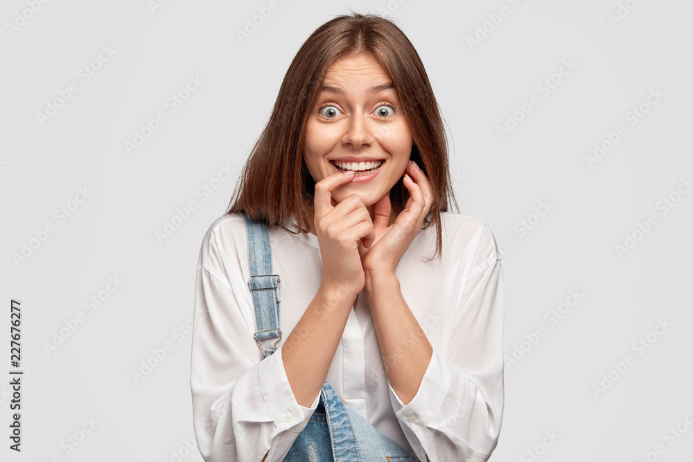 Beautiful teenage girl feels happy, smiles broadly, keeps index finger near mouth, looks curiously at camera, dressed in fashionable clothes, isolated over white background. Positive emotions concept Beautiful teenage girl feels happy, smiles broadly, keeps index finger near mouth, looks curiously at camera, dressed in fashionable clothes, isolated over white background. Positive emotions concept