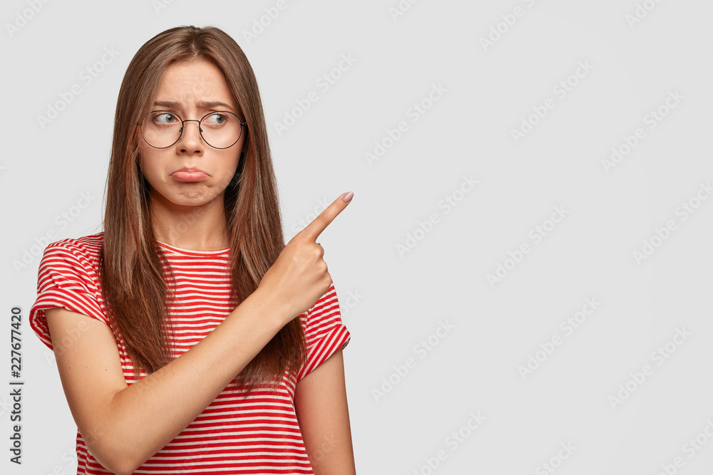 Horizontal view of dejected displeased dark haired woman purses lips with dissatisfaction, has some troubles, points with index finger at upper right corner, doesnt like something. Negative emotions Horizontal view of dejected displeased dark haired woman purses lips with dissatisfaction, has some troubles, points with index finger at upper right corner, doesnt like something. Negative emotions