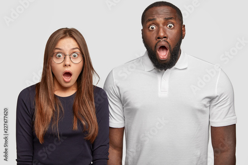 Fotografi Surprised dark skinned adult man and emotive stupefied Caucasian woman keep jaws