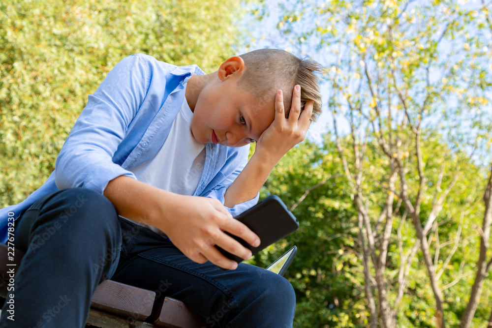 childhood, augmented reality, technology and people concept - boy with upset face looks into the smartphone outdoors at summer childhood, augmented reality, technology and people concept - boy with upset face looks into the smartphone outdoors at summer