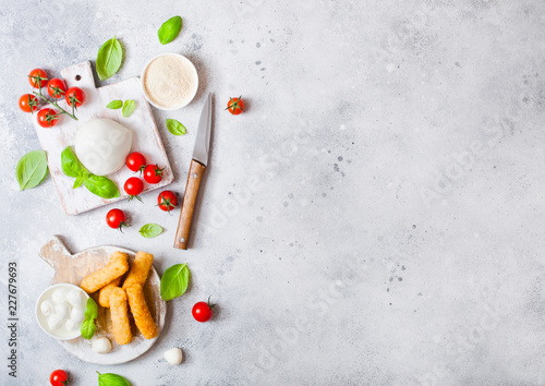 Fresh Mozzarella cheese on vintage chopping board with tomatoes and basil leaf and tray with cheese sticks on stone kitchen table background. Space for text