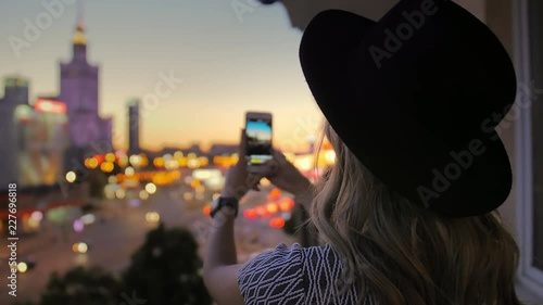 The girl in the hat makes a photo of the night city. Uses a smartphone. In the frame of a skyscraper in the center of the capital. Warsaw.