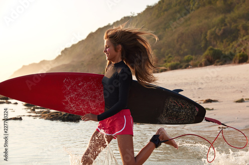 View of active sporty woman runs on water in warm ocean, during windy weather, carries surfboard hurries to hit waves, being photographed in motion against rocky cliff. Real paradise place for rest photo