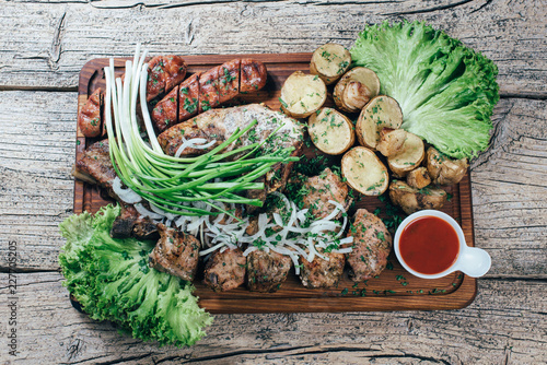 Appetizing roasted pork pieces on the grill, presented on a wooden board, along with leaves of green salad and potatoes with tomato sauce