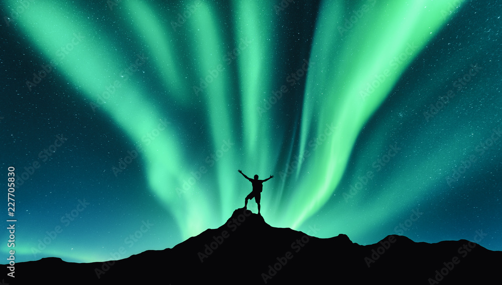 Northern lights and silhouette of standing man with raised up arms on the mountain in Norway. Aurora borealis and happy man. Sky with stars and green polar lights. Night landscape with aurora. Concept - obrazy, fototapety, plakaty 