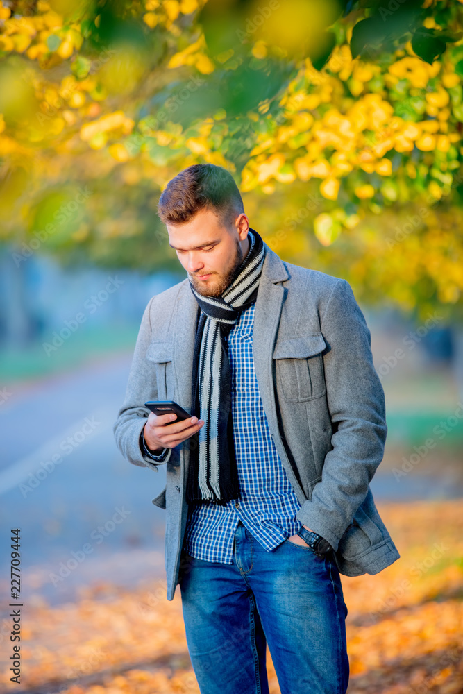 Fototapeta premium Young man with mobile phone in autumn season alley.