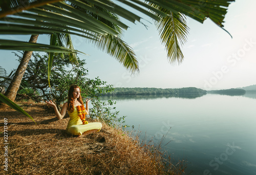 A woman is meditating by the river with a garland of flowers around her neck. In the foreground hangs a palm leaf from above.
