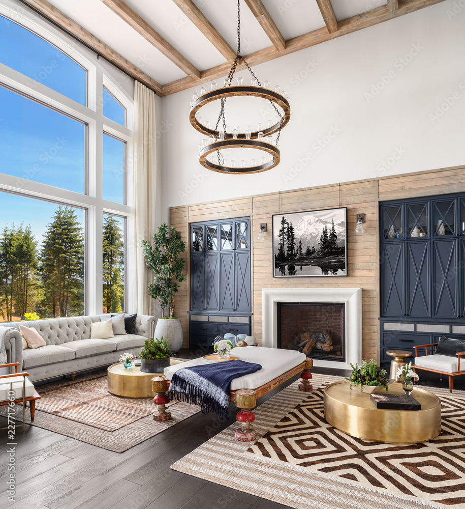 Beautiful Living Room in Luxury Home with Elegant Chandelier, Fireplace, Hardwood Floors, and Tall Ceiling. Has Large Bank of Windows with Exterior View of Blue Sky and Trees. - obrazy, fototapety, plakaty 