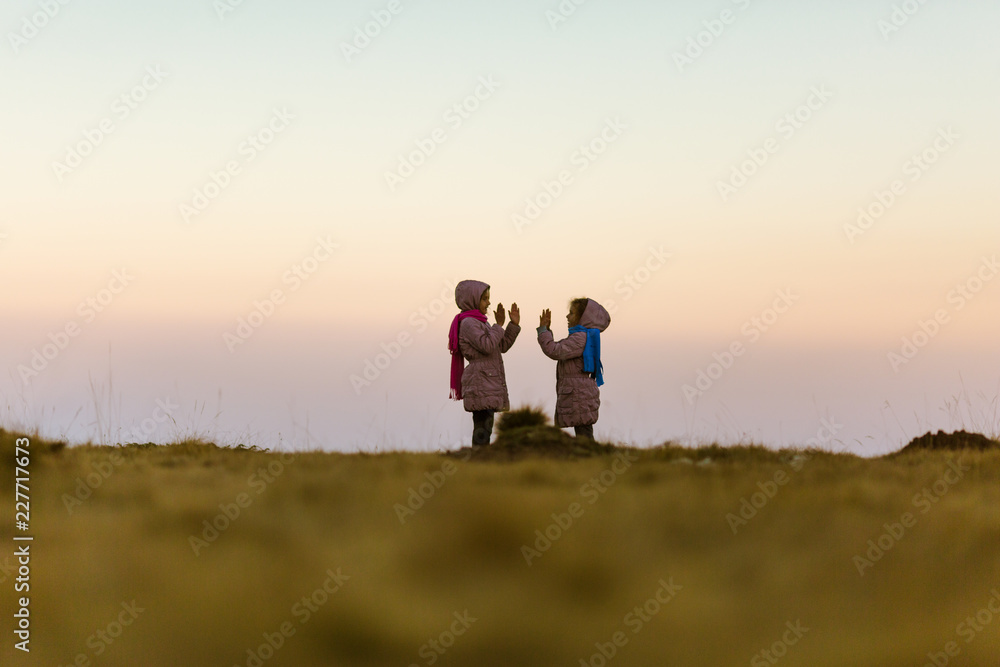 Two young girls in nature, playing clapping handgame. Girls are 7 years old and are twin sisters. Dressed in pink winter coats with shawls. Beautiful blurred background. Dusk. Two young girls in nature, playing clapping handgame. Girls are 7 years old and are twin sisters. Dressed in pink winter coats with shawls. Beautiful blurred background. Dusk.