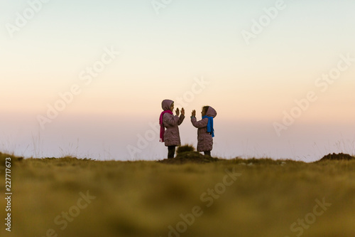 Two young girls in nature, playing clapping handgame. Girls are 7 years old and are twin sisters. Dressed in pink winter coats with shawls. Beautiful blurred background. Dusk.
