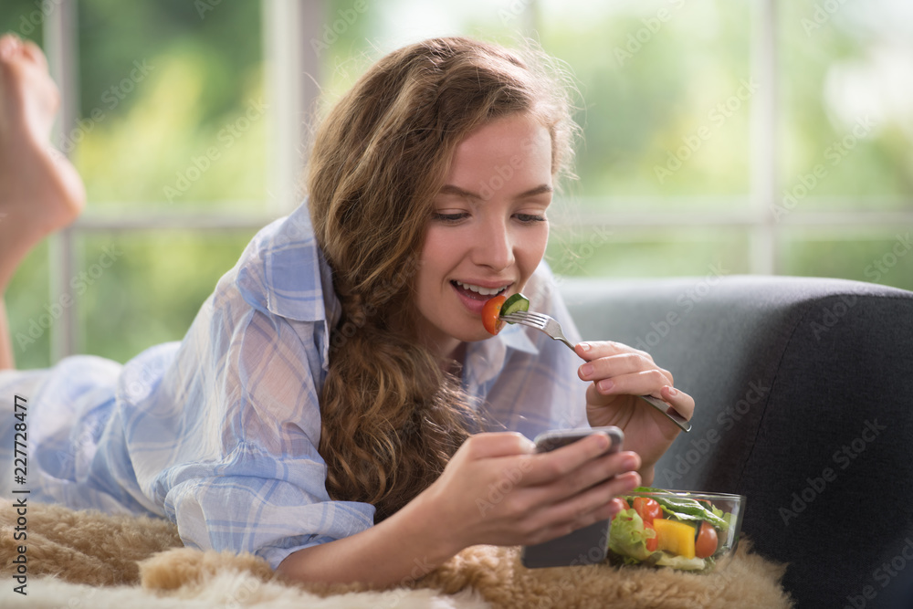 Healthy young woman lying on a couch holding a salad bowl looking relaxed and comfortable Healthy young woman lying on a couch holding a salad bowl looking relaxed and comfortable