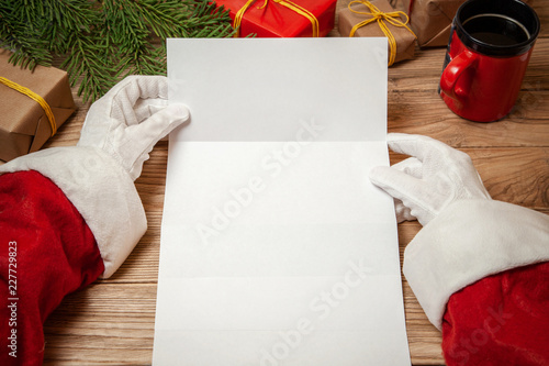 Santa Claus holding letter on wooden table with gift boxes and Christmas tree and cup of hot coffee or tea.  mockup blank