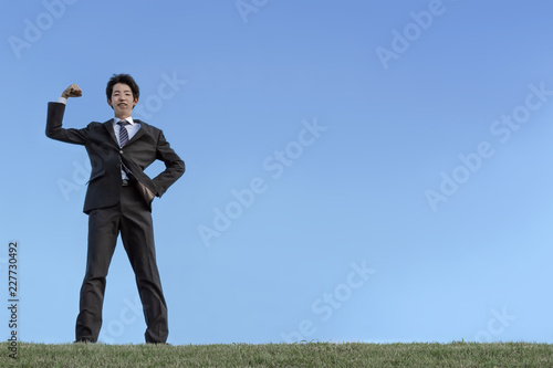 青空をバックにガッツポーズをするスーツ姿の若いビジネスマン1人。元気、成功、ヤル気、挑戦イメージ
