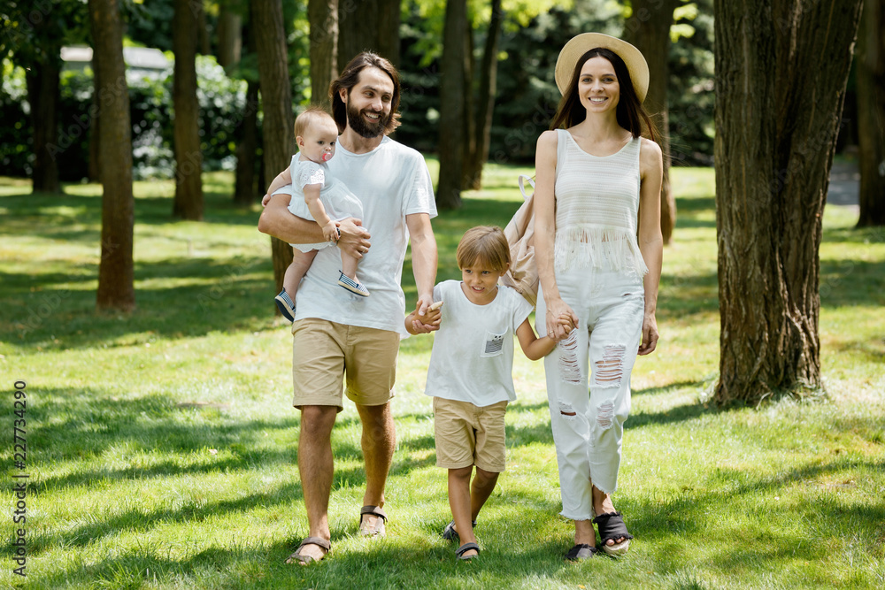 Warm sunny day. Handsome father, beautiful mother and two kids dressed in the white casual clothes happily walks in the park. Warm sunny day. Handsome father, beautiful mother and two kids dressed in the white casual clothes happily walks in the park.