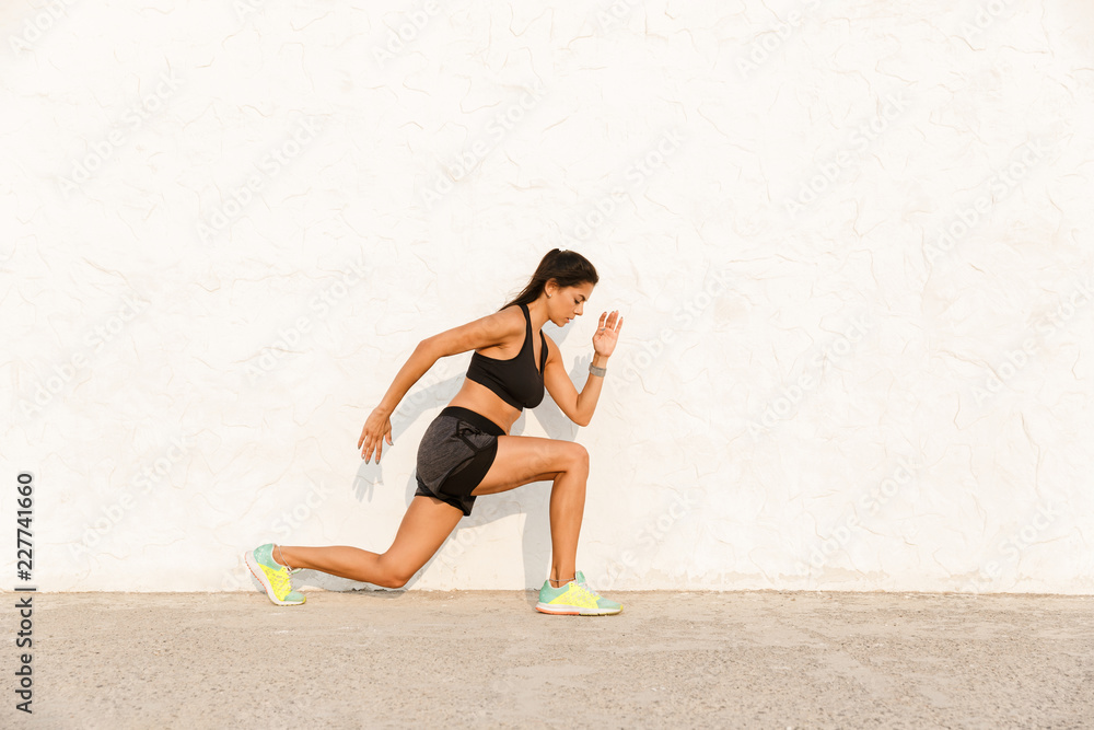 Full length image of brunette girl 20s in tracksuit squatting and stretching her body, over wall Full length image of brunette girl 20s in tracksuit squatting and stretching her body, over wall