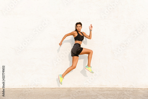 Full length image of beautiful woman 20s in sportswear working out and running, along wall