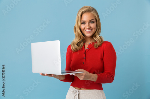 Portrait of charming blond woman 20s holding silver laptop, isolated over blue background in studio